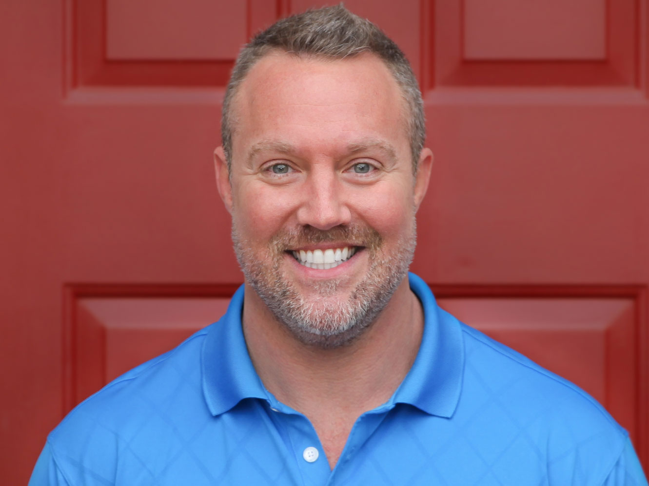 Grant Golliver, owner of Messick Roofing, smiling in a blue polo in front of a red door.